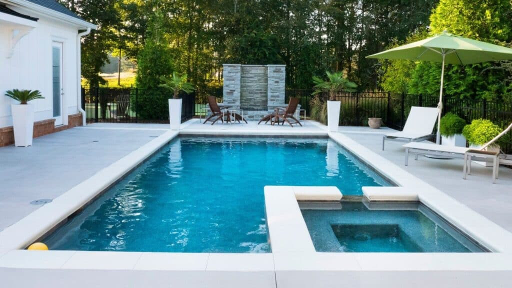Modern backyard with a rectangular swimming pool and attached hot tub, surrounded by lounge chairs, an umbrella, potted plants, and a small dining set. Tall green trees and a privacy fence are in the background.