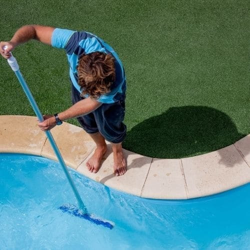 A person standing barefoot at the edge of a swimming pool uses a pole brush to clean the pool, with artificial grass visible in the background.