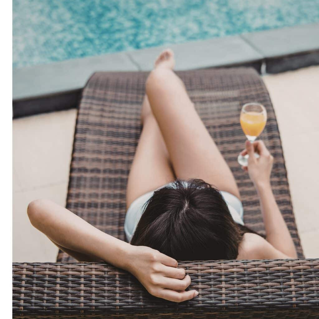 A woman in a swimsuit relaxes on a woven lounge chair by a sparkling swimming pool, freshly maintained with expert pool cleaning from Inman Pool Service, holding a glass of orange beverage, viewed from above.