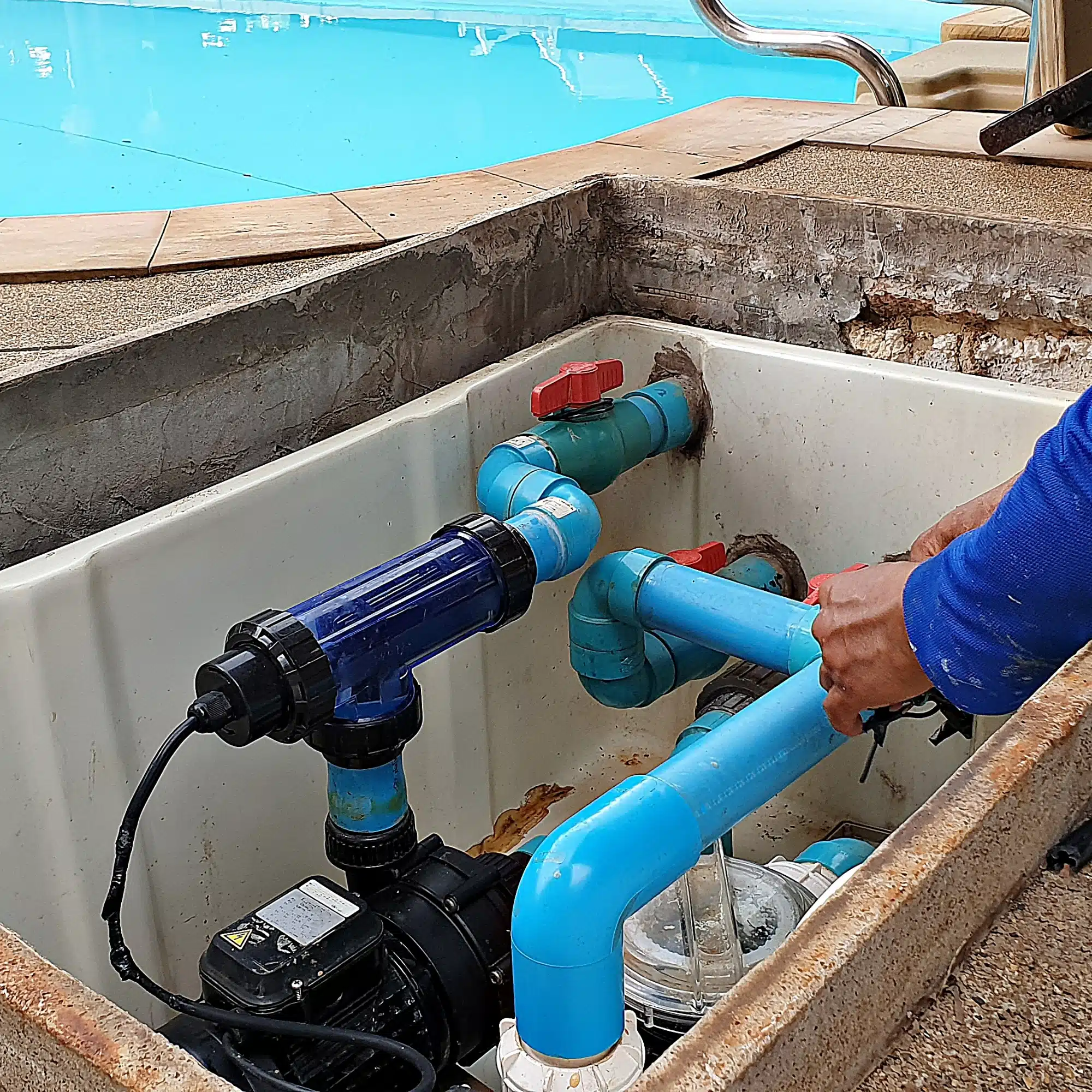 A person in a blue shirt from Inman Pool Service is working on blue PVC pool plumbing and filtration equipment near a swimming pool, adjusting pipes and connections in an open maintenance compartment.