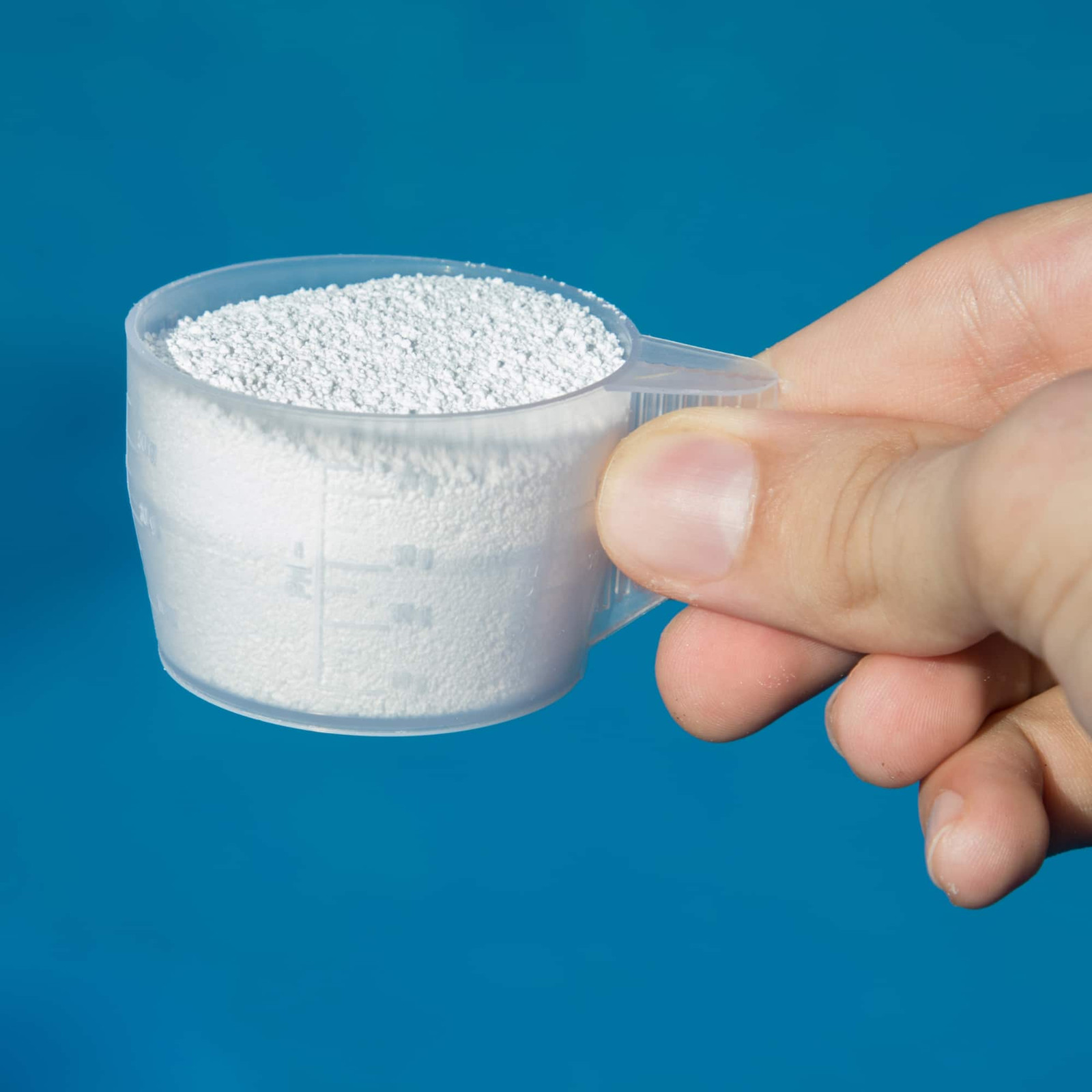 A hand holds a small, clear plastic scoop filled with white granular powder against a blue background—perfect for pool maintenance with Inman Pool Service.