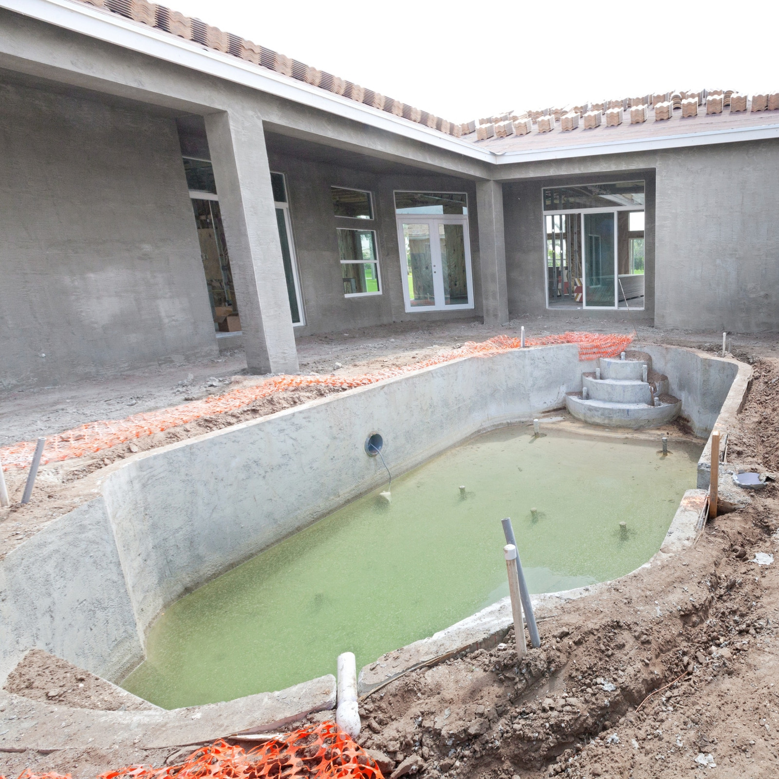 A partially constructed, empty swimming pool with some greenish water at the bottom—an ideal candidate for Inman Pool Service’s expert pool cleaning—is surrounded by dirt and orange safety barriers, next to an unfinished house with bare walls and exposed windows.