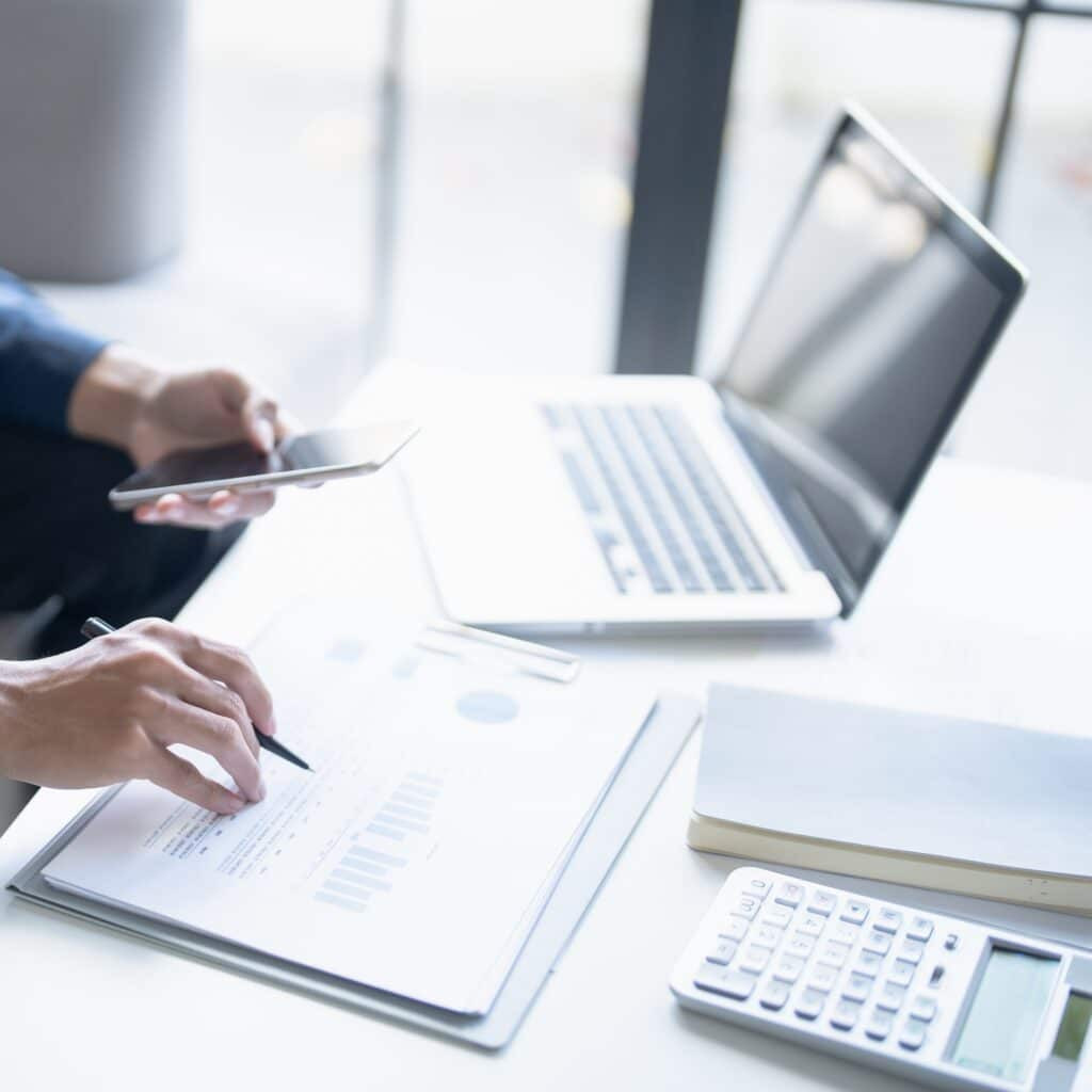 A person works at a desk with a laptop, smartphone, printed charts, pen, calculator, and notebook—analyzing documents and taking pool service calls for Inman. The scene is bright with natural light from a nearby window.