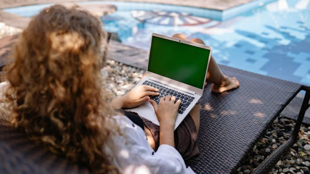A person with curly hair sits on a lounge chair by a pool in the service area, working on a laptop with a green screen. Pebbles and water are visible nearby, creating a relaxed outdoor atmosphere.