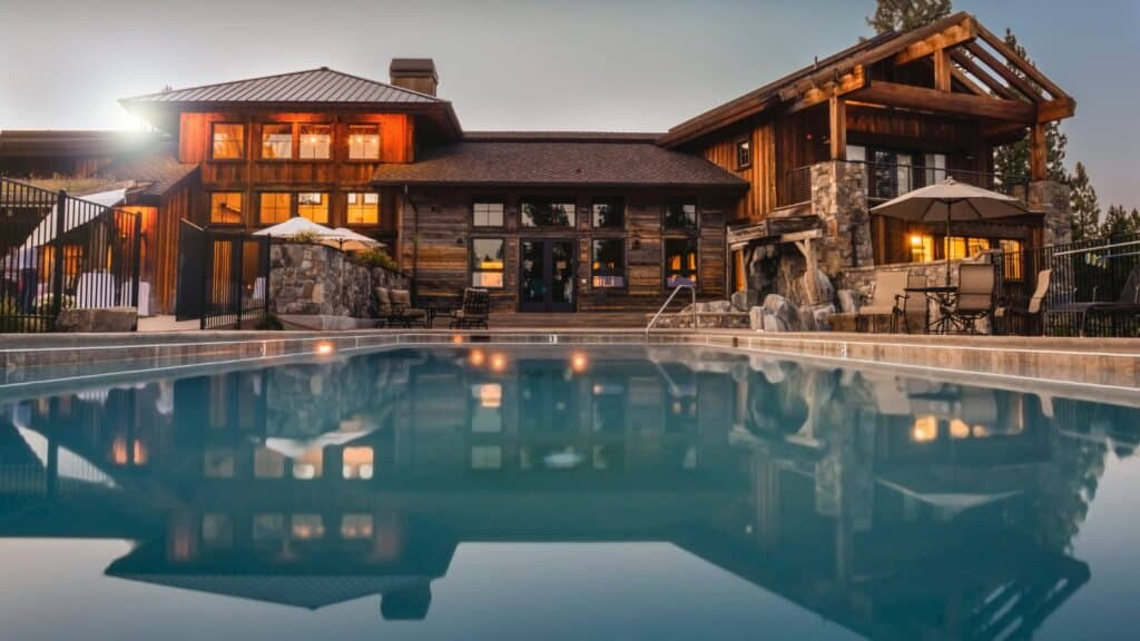 A large, modern wooden house with glowing lights is reflected in a clear swimming pool at dusk, surrounded by outdoor seating and umbrellas.