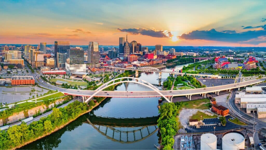 Aerial view of downtown Nashville, Tennessee at sunset, highlighting the John Seigenthaler Pedestrian Bridge spanning the Cumberland River within this vibrant service area, with modern buildings and a colorful sky.